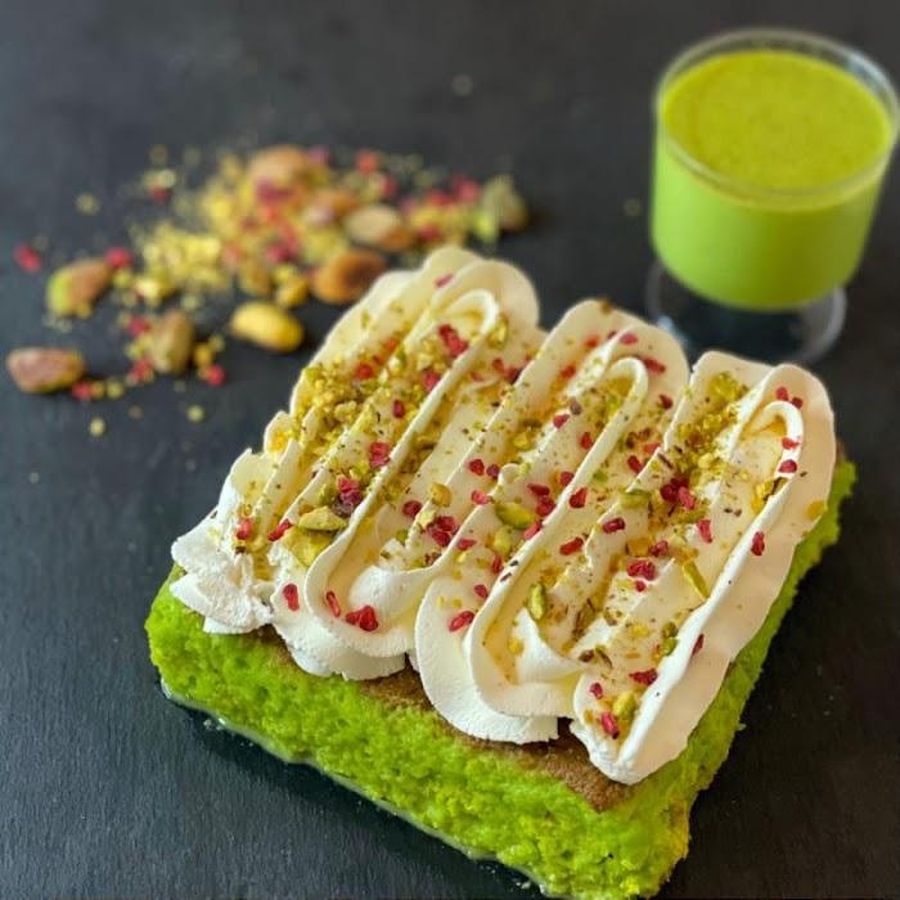 A slice of pistachio milk cake topped with fresh cream, pistachio crumb and a rose sprinkle, with a pistachio milkshake alongside at Cakes & Craves